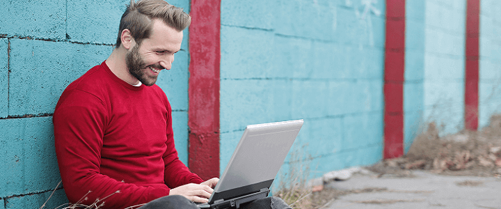 stock photo of man sitting outside against blue cinderblock wall with computer