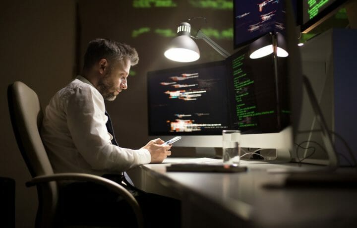 Man sitting at desk in the dark on phone with computers and code