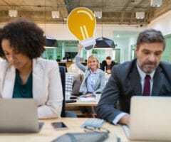 Woman in background of office holding giant lightbulb over head