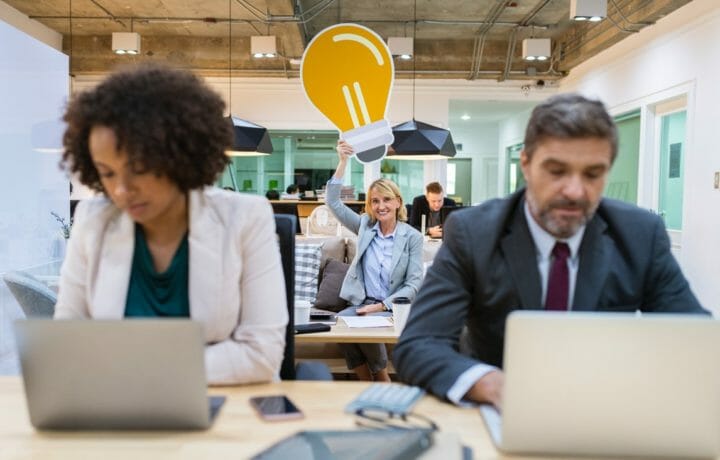 Woman in background of office holding giant lightbulb over head