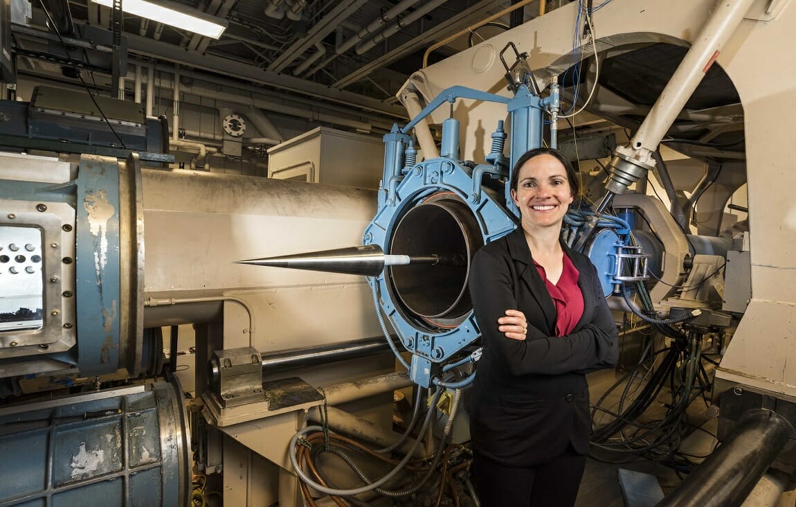 slender woman in black clothes and pink shirt smiling standing in front of aerospace equipment