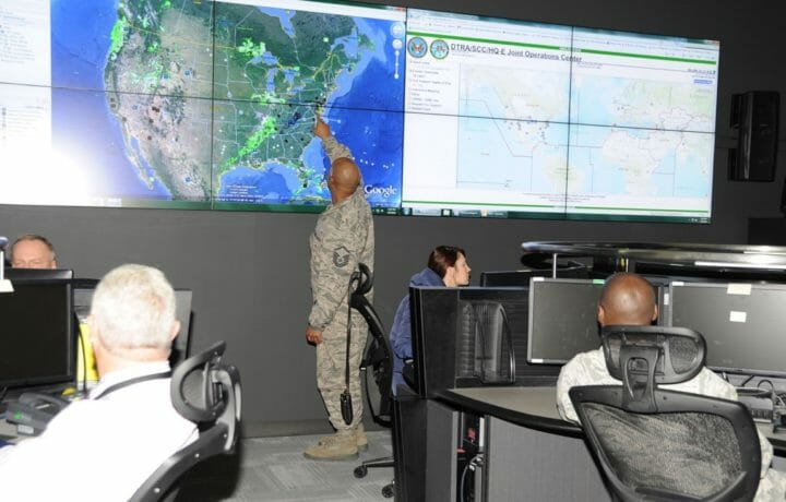 man in uniform pointing to a screen with a map in an office with other workers at desks in the defense threat reduction agency