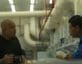 man with tattoos and camera standing in factory next to man in latex gloves Northrop Grumman