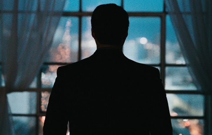 silhouette of a mysterious man in front of a window looking at a city at nighttime