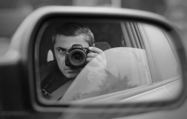 man in driver's seat of a car holding camera spying on someone