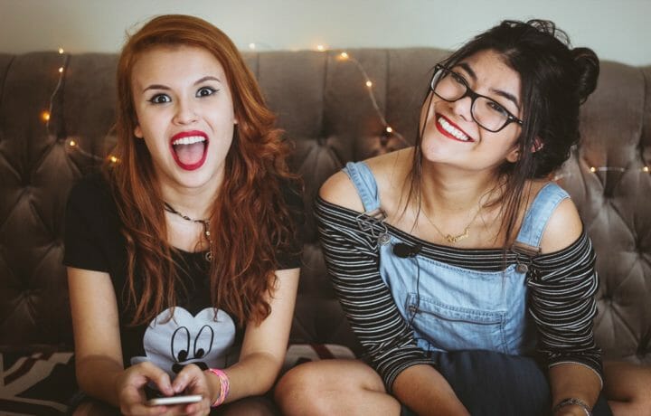 teenage girl with red hair and red lipstick and goofy face sitting next to a dark haired teenage girl with glasses and overalls smiling