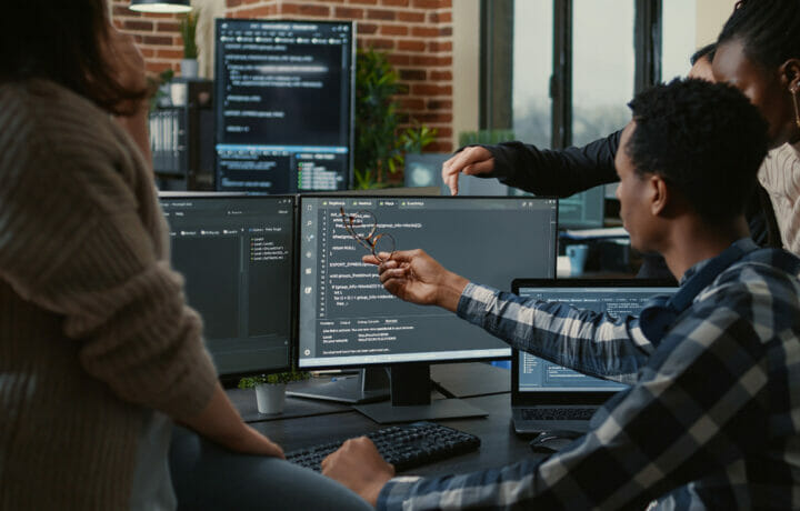 Software developers discussing about source code compiling discovers errors and asks the rest of the team for explanations in front of multiple screens running algorithms. Programmers doing teamwork.