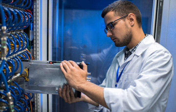 Side view portrait of young man in lab coat taking out blade server out of cabinet while working with supercomputer