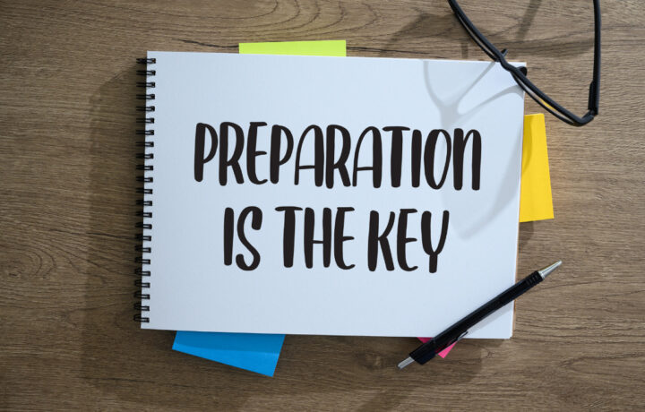 "Preparation is the key" written on a notebook with post-its sticking out. A pair of glasses and a pen are on the notebook.