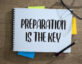 "Preparation is the key" written on a notebook with post-its sticking out. A pair of glasses and a pen are on the notebook.