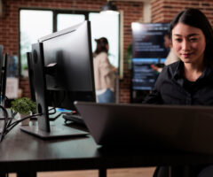 Image of woman smiling as she looks at a computer. There are computers and people in the background, it looks like an office building.