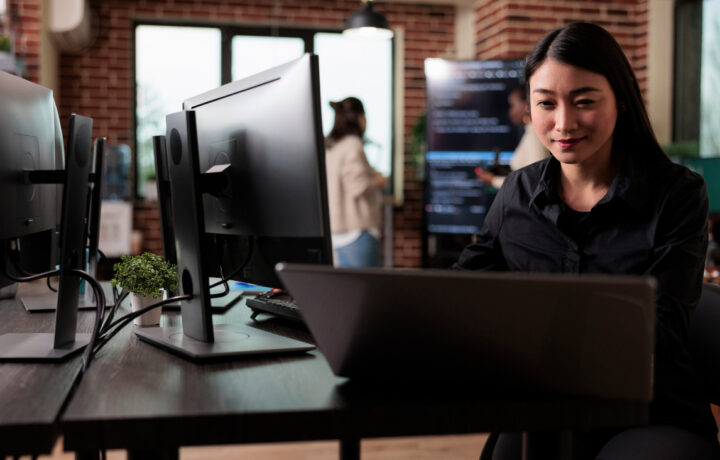 Image of woman smiling as she looks at a computer. There are computers and people in the background, it looks like an office building.