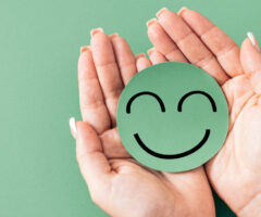 Photo of manicured hands holding a green smiley face, against the same color green background.