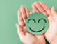 Photo of manicured hands holding a green smiley face, against the same color green background.