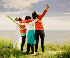 Four women with their backs to the camera, arms around each other facing the ocean and sky.