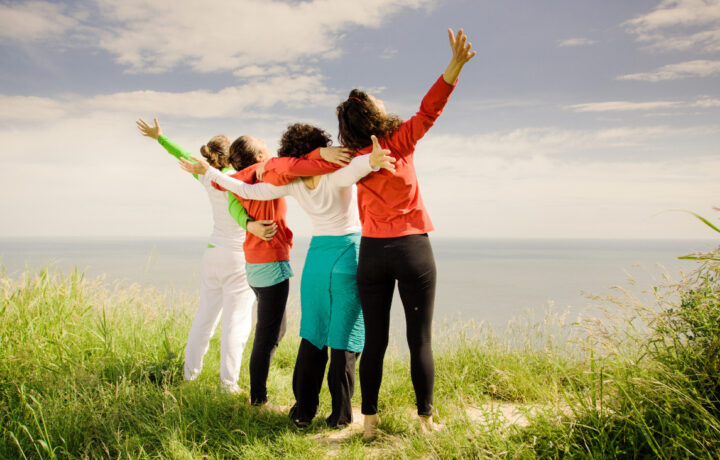 Four women with their backs to the camera, arms around each other facing the ocean and sky.