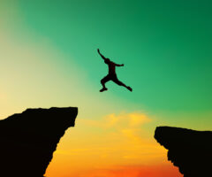 Photo of someone jumping between cliffs. The cliffs and person in the air between them are black while the sky in the background is blue/green on top and yellow and orange on the bottom.