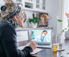 Photo of a woman sitting at a kitchen table talking to a doctor on the laptop.