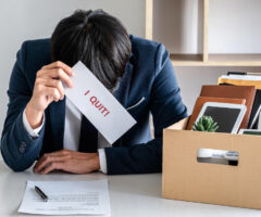 A frustrated employee in a business suit sits at a desk holding a resignation letter labeled "I QUIT!" in one hand, with a packed cardboard box containing personal belongings, including a clock and office supplies, in the background.