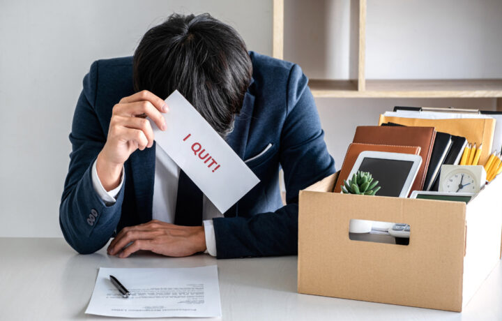 A frustrated employee in a business suit sits at a desk holding a resignation letter labeled "I QUIT!" in one hand, with a packed cardboard box containing personal belongings, including a clock and office supplies, in the background.