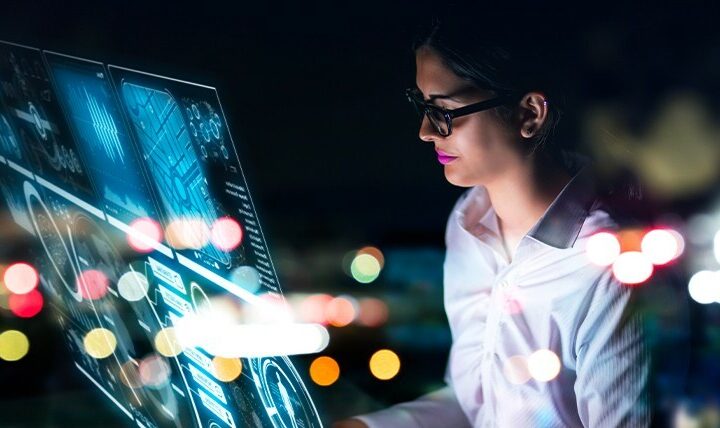 A woman wearing glasses and a white shirt is working on a futuristic, high-tech digital interface displaying data visualizations and analytics. The scene is illuminated by the glow of the screen, with blurred city lights in the background, creating a modern and technology-focused atmosphere.