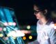 A woman wearing glasses and a white shirt is working on a futuristic, high-tech digital interface displaying data visualizations and analytics. The scene is illuminated by the glow of the screen, with blurred city lights in the background, creating a modern and technology-focused atmosphere.