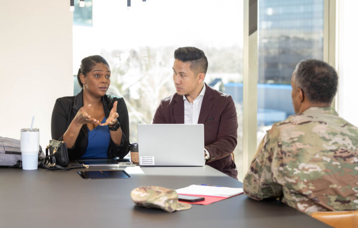 A professional meeting takes place in a modern office with large windows allowing natural light. A woman in a black blazer and blue blouse gestures as she speaks, while a man in a maroon blazer listens attentively, with a laptop open in front of him. Across from them, a person in a military uniform sits with their back to the camera. A camouflage cap, notepad, and smartphone are placed on the table, alongside personal belongings like a backpack and a tumbler.