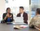 A professional meeting takes place in a modern office with large windows allowing natural light. A woman in a black blazer and blue blouse gestures as she speaks, while a man in a maroon blazer listens attentively, with a laptop open in front of him. Across from them, a person in a military uniform sits with their back to the camera. A camouflage cap, notepad, and smartphone are placed on the table, alongside personal belongings like a backpack and a tumbler.