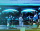 A high-tech operations center with multiple computer workstations and large screens displaying a digital rendering of Earth. A diverse team of cybersecurity or intelligence professionals, dressed in business casual attire, is engaged in discussion and analysis. The image has a futuristic overlay of binary code (1s and 0s), symbolizing data processing and cyber intelligence.