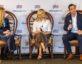 Three panelists sit on a stage during a ClearanceJobs event, participating in a discussion. Behind them is a branded backdrop with the ClearanceJobs logo. The woman on the left wears a navy pantsuit and holds a microphone. The woman in the center wears a white blouse and dark wide-leg trousers, also holding a microphone. The man on the right wears a navy suit and is mid-sentence, gesturing with one hand while holding a microphone in the other. All three are seated on wooden chairs, engaged in conversation.