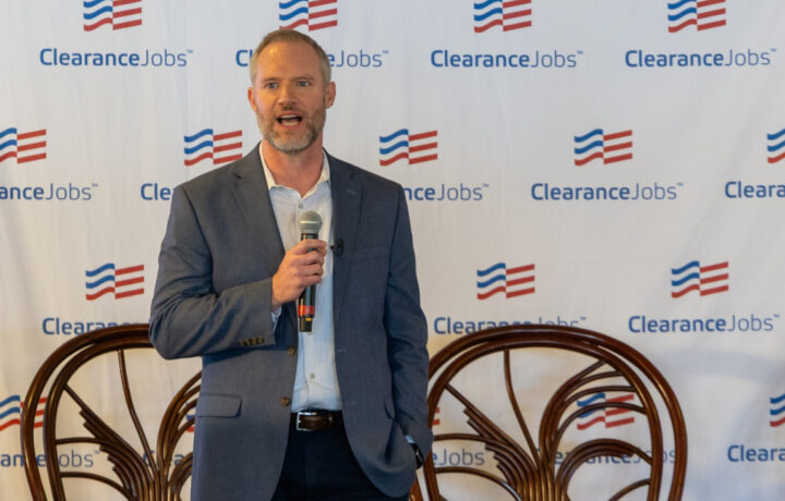 Alex Schildt, President of ClearanceJobs, speaks on stage holding a microphone at ClearanceJobs Connect West, with a branded backdrop behind him.