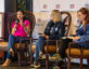 Three women sit on a panel stage at ClearanceJobs Connect 2025 West in San Diego, California. The speaker on the left, wearing a bright pink top, gestures while speaking into a microphone. The center panelist wears a dark hoodie with a colorful unicorn logo and holds a mic. The moderator on the right, wearing a blue velvet jacket, listens attentively with notes in hand. A branded ClearanceJobs backdrop and event signage are visible behind them.