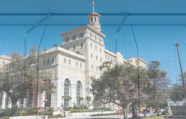 A large, historic building with a prominent clock tower and a cross on top is visible. The structure features a light-colored facade with multiple windows and decorative elements. In front of the building, there are trees and landscaped areas. A grid-like mesh overlay partially obscures the view, creating a layered effect. The sky is clear and blue, enhancing the overall brightness of the scene.