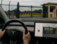iew from inside a Tesla with hands on the steering wheel, approaching a secure government facility gate with barbed-wire fencing, a guardhouse, and a stop sign visible through the windshield.