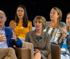 Five people engaged in a discussion, seated in a row. From left to right: a man in a blue blazer and glasses gesturing with his hands; a woman in a mustard yellow sleeveless top speaking; a woman in a gray suit with short blonde hair listening attentively; a woman in a patterned top and green cardigan holding a microphone; and a woman with pink-streaked hair in a gray blazer and blue shirt also holding a microphone and gesturing.