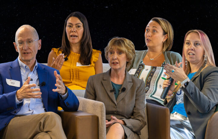 Five people engaged in a discussion, seated in a row. From left to right: a man in a blue blazer and glasses gesturing with his hands; a woman in a mustard yellow sleeveless top speaking; a woman in a gray suit with short blonde hair listening attentively; a woman in a patterned top and green cardigan holding a microphone; and a woman with pink-streaked hair in a gray blazer and blue shirt also holding a microphone and gesturing.