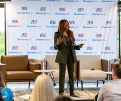 A woman in a dark green suit is standing and speaking on a stage in front of an audience. She holds a tablet in one hand and gestures with the other. Behind her is a backdrop with the "ClearanceJobs" logo repeated across it. The stage has several chairs and small tables, and a flag is visible on the right side. The audience is seated and facing the speaker.