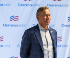 A middle-aged man with light brown hair, wearing a blue checkered blazer over a light blue dress shirt, stands in front of a backdrop featuring the "ClearanceJobs" logo repeatedly printed in blue and red. The man has a serious expression and is looking slightly to his right.