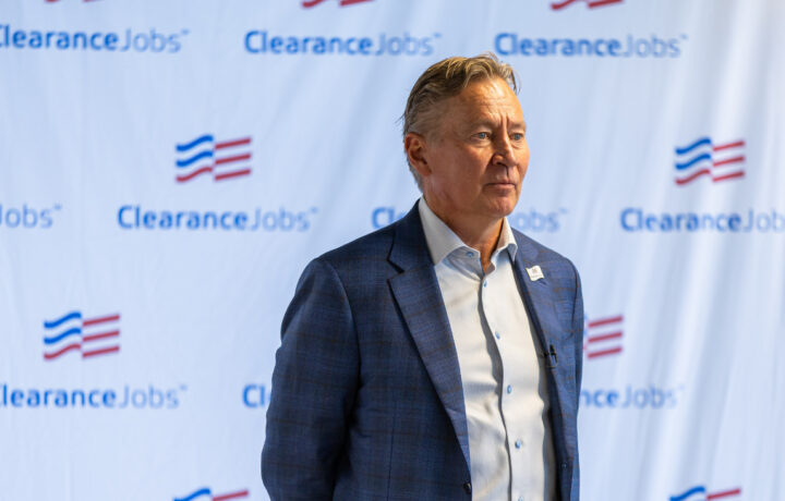 A middle-aged man with light brown hair, wearing a blue checkered blazer over a light blue dress shirt, stands in front of a backdrop featuring the "ClearanceJobs" logo repeatedly printed in blue and red. The man has a serious expression and is looking slightly to his right.