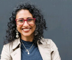 A smiling woman with curly black hair wearing red-framed glasses, large round earrings, a beige trench coat, a dark blue top, and layered silver necklaces, standing against a plain dark gray background.