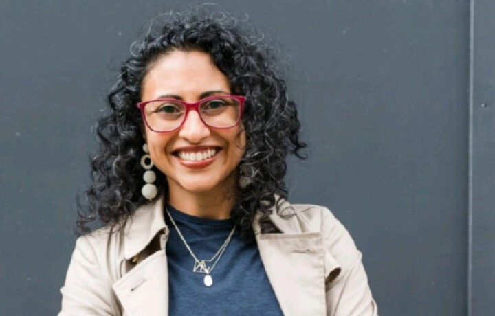 A smiling woman with curly black hair wearing red-framed glasses, large round earrings, a beige trench coat, a dark blue top, and layered silver necklaces, standing against a plain dark gray background.