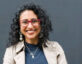 A smiling woman with curly black hair wearing red-framed glasses, large round earrings, a beige trench coat, a dark blue top, and layered silver necklaces, standing against a plain dark gray background.
