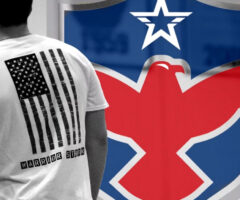 A person wearing a white T-shirt with a distressed black American flag design on the back. Below the flag, the text "WARRIOR STRON" is printed in black block letters. In the background, there is a large emblem featuring a red eagle with outstretched wings on a blue shield, topped with a white star.