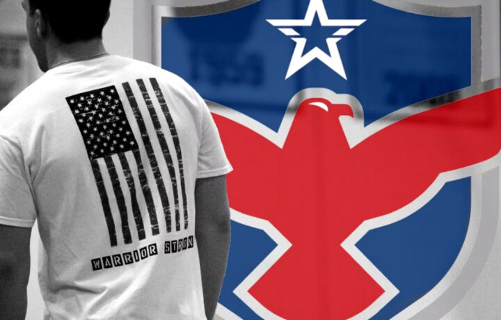 A person wearing a white T-shirt with a distressed black American flag design on the back. Below the flag, the text "WARRIOR STRON" is printed in black block letters. In the background, there is a large emblem featuring a red eagle with outstretched wings on a blue shield, topped with a white star.