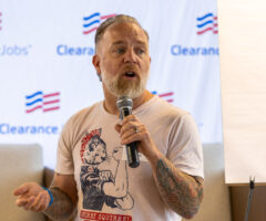A man with a beard and tattoos on his left arm is speaking into a microphone. He is wearing a light-colored T-shirt featuring an illustration of a squirrel dressed like Rosie the Riveter with the text "SECRET SQUIRREL." The background shows a banner with the "ClearanceJobs" logo and a blank flip chart.