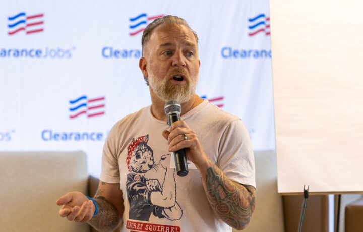 A man with a beard and tattoos on his left arm is speaking into a microphone. He is wearing a light-colored T-shirt featuring an illustration of a squirrel dressed like Rosie the Riveter with the text "SECRET SQUIRREL." The background shows a banner with the "ClearanceJobs" logo and a blank flip chart.