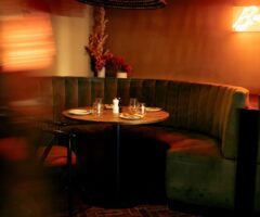 Curved green velvet booth seating around a round wooden table set with plates, glasses, and salt and pepper shakers, in a dimly lit restaurant with warm orange lighting and a neon sign on the wall.
