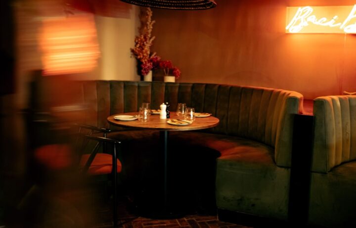 Curved green velvet booth seating around a round wooden table set with plates, glasses, and salt and pepper shakers, in a dimly lit restaurant with warm orange lighting and a neon sign on the wall.