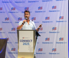 A man with short brown hair and a beard is speaking into a microphone while standing behind a podium. The podium has a sign that reads "ClearanceJobs CONNECT 2025" with a small squirrel logo. The background features a large banner with repeated "ClearanceJobs" logos and red, white, and blue flag-like symbols. To the right, there is a display board with various logos and text, partially visible. A dark brown chair is positioned to the left of the podium.