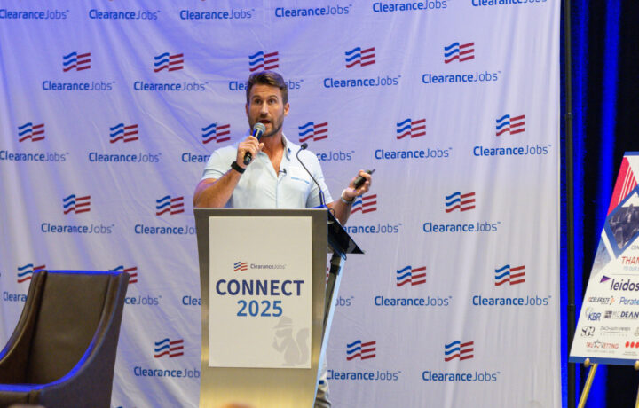 A man with short brown hair and a beard is speaking into a microphone while standing behind a podium. The podium has a sign that reads "ClearanceJobs CONNECT 2025" with a small squirrel logo. The background features a large banner with repeated "ClearanceJobs" logos and red, white, and blue flag-like symbols. To the right, there is a display board with various logos and text, partially visible. A dark brown chair is positioned to the left of the podium.
